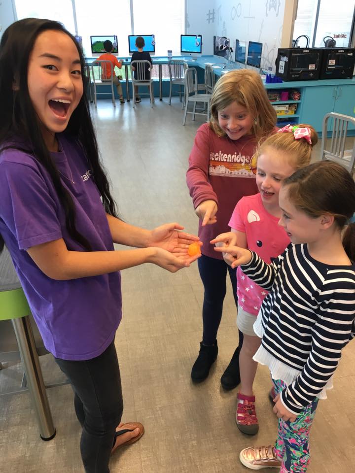 A smiling woman holds a small orange object while three excited young girls point at it. They are in a bright room with computers and chairs in the background.