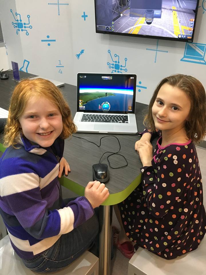 Two smiling children sit at a table, looking at the camera while playing a space simulation game on a laptop. Science-themed drawings and a large monitor are visible in the background.
