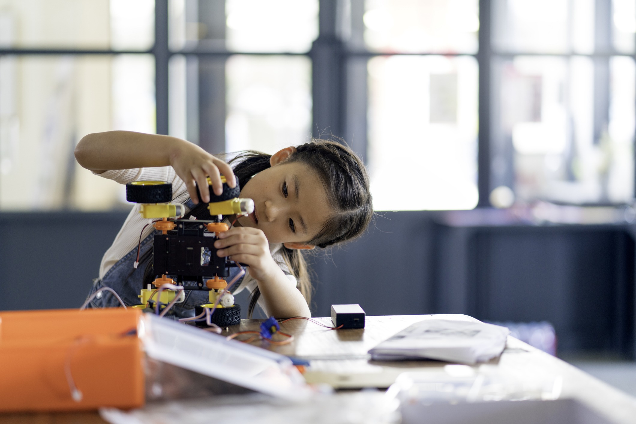 A young girl carefully assembles a small robotic vehicle at a table, surrounded by tools and papers, in a bright room with large windows in the background.