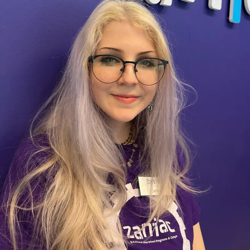A young person with long, light blonde hair, wearing round glasses and a purple t-shirt stands in front of a purple wall. The t-shirt has a white circular logo and they are smiling at the camera.