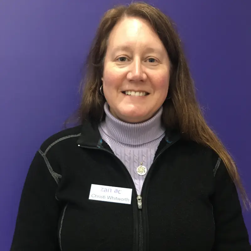 A woman with long brown hair, wearing a black jacket over a lavender turtleneck, smiles in front of a solid purple background. She has a name tag on her jacket.