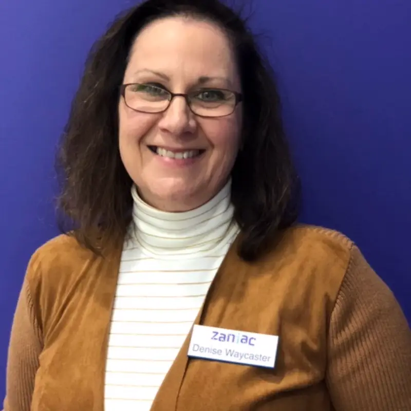 A woman with shoulder-length brown hair, glasses, and a white turtleneck under a brown vest smiles in front of a solid blue background. She is wearing a name tag that reads "zaniac Denise Waycaster.