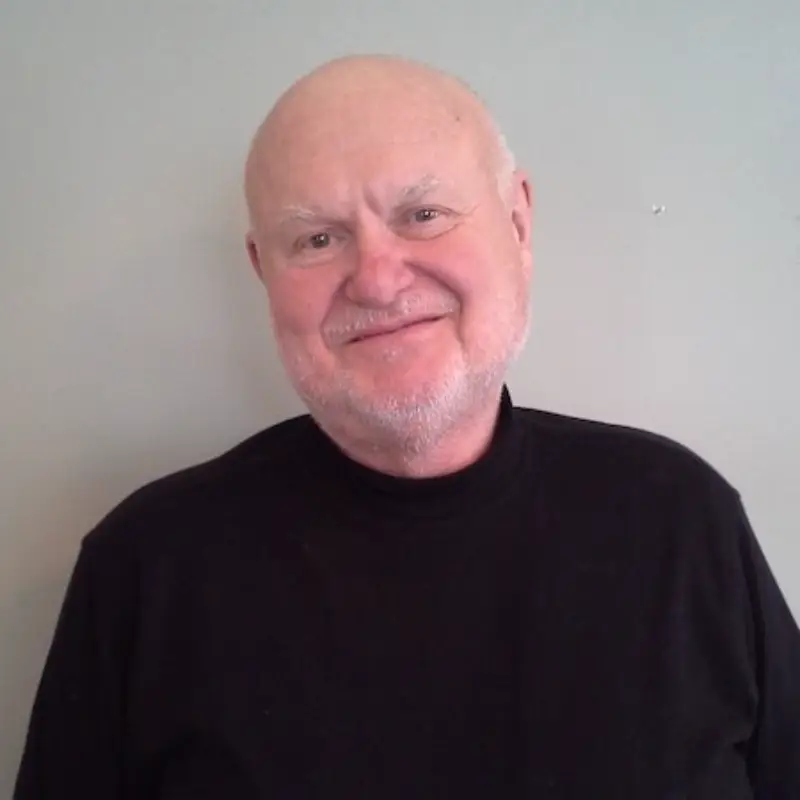 A smiling older man with a bald head and white beard, wearing a black turtleneck sweater, standing against a plain light gray wall.