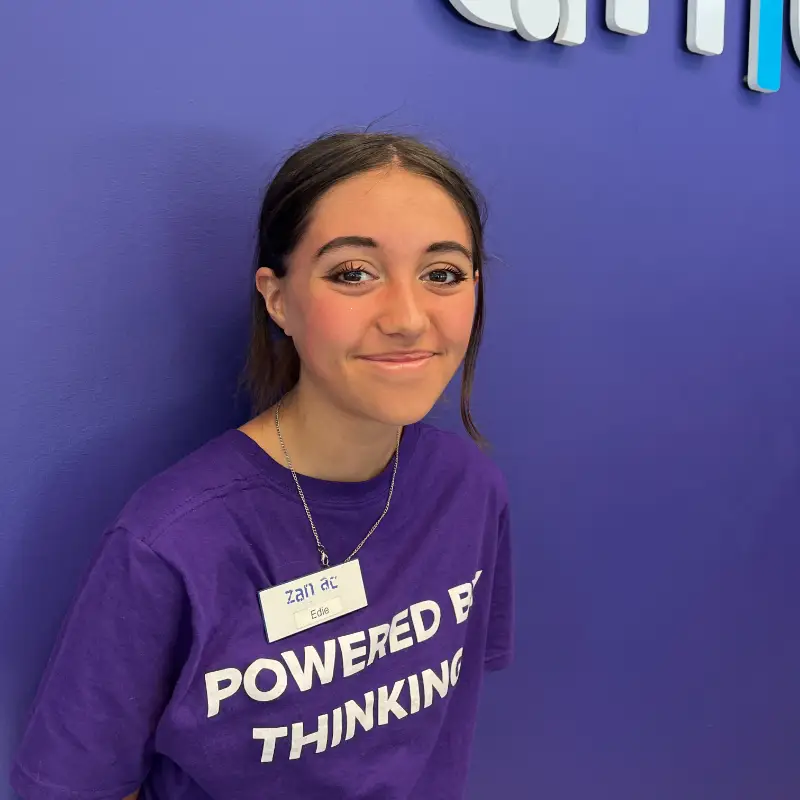A person with long brown hair wearing a purple "Powered by Thinking" t-shirt and a name tag stands smiling in front of a blue wall with part of a logo visible.