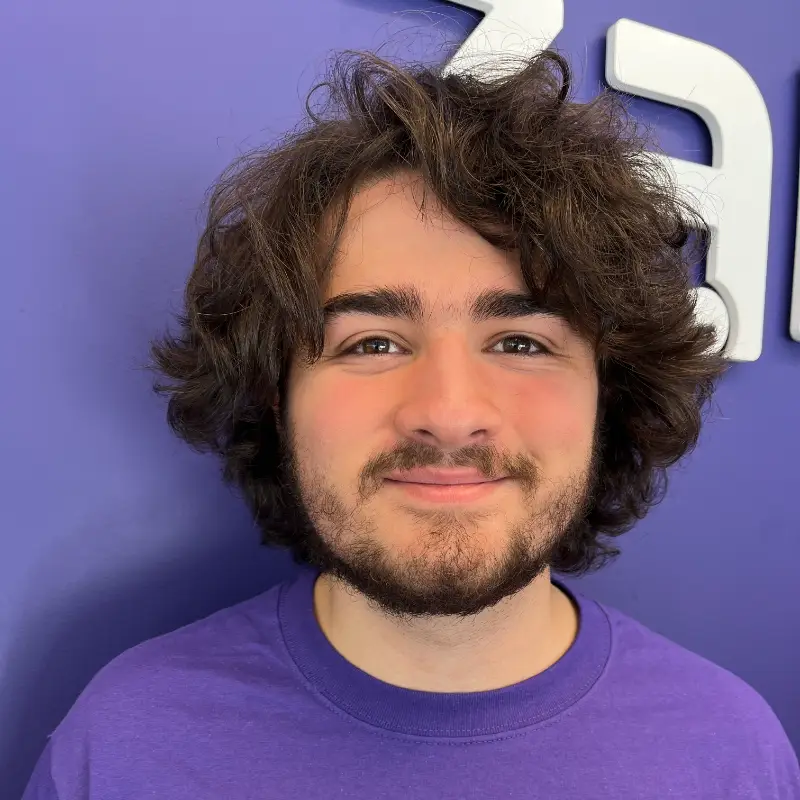 A young person with wavy brown hair and a beard smiles at the camera, wearing a purple shirt, standing in front of a purple wall with part of a white logo visible.