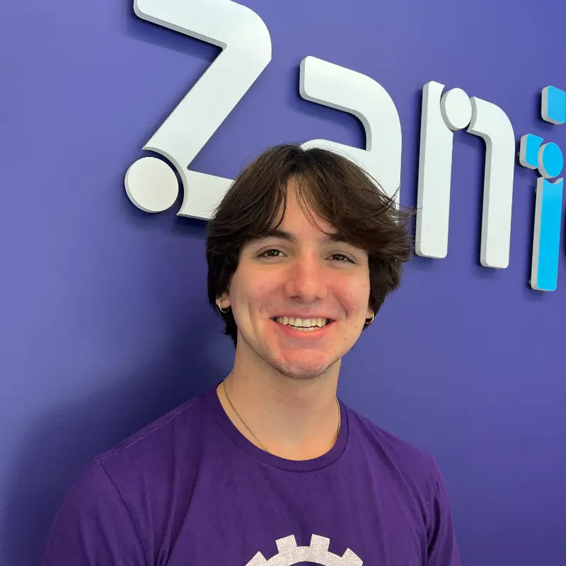 A young person with medium-length brown hair and a purple shirt smiles while standing in front of a blue wall with large white and blue letters that partially spell "Zani.