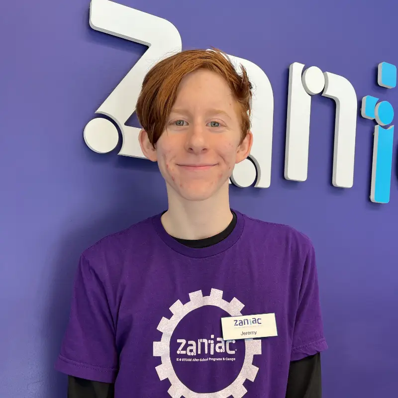 A smiling youth with red hair stands in front of a blue wall with "zaniac" written on it. They are wearing a purple Zaniac T-shirt and a name tag that says "Jeremy.