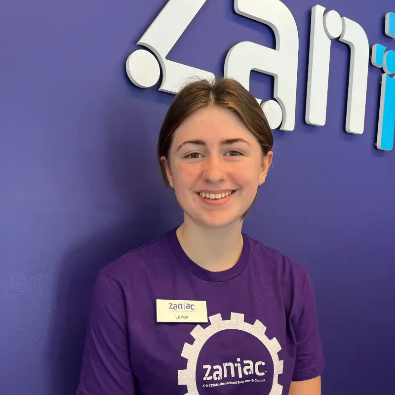 A young woman with brown hair, wearing a purple Zaniac shirt and a name tag that says "Laney," smiles while standing in front of a purple wall with the Zaniac logo.