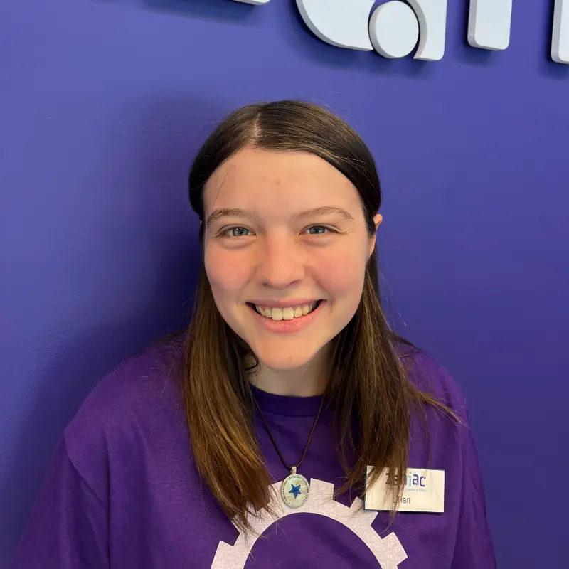 A young person with straight brown hair, wearing a purple shirt and a necklace with a blue star pendant, smiles in front of a blue wall. They have a name tag on their shirt.