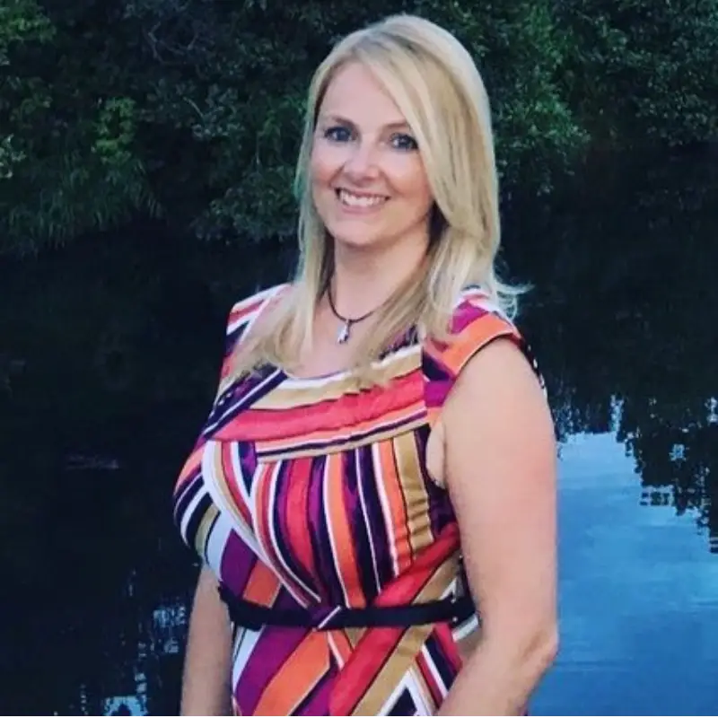 A smiling woman with long blonde hair stands outdoors by water, wearing a colorful, striped sleeveless dress. Green foliage is visible in the background.