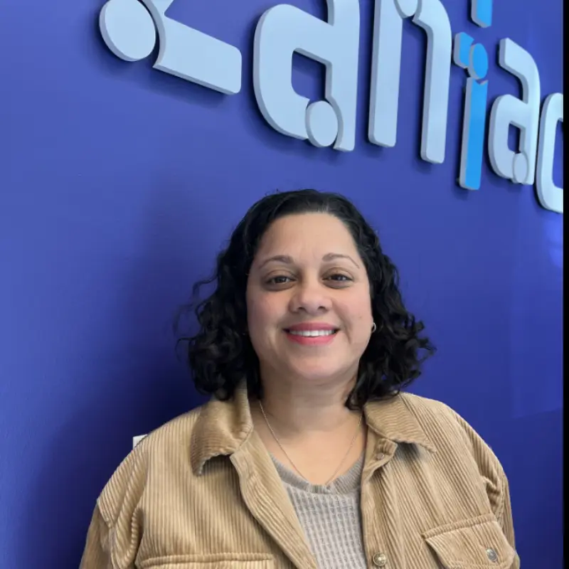 A woman with curly dark hair and a tan corduroy jacket smiles in front of a blue wall with a large white and blue logo partially reading "an" in the background.
