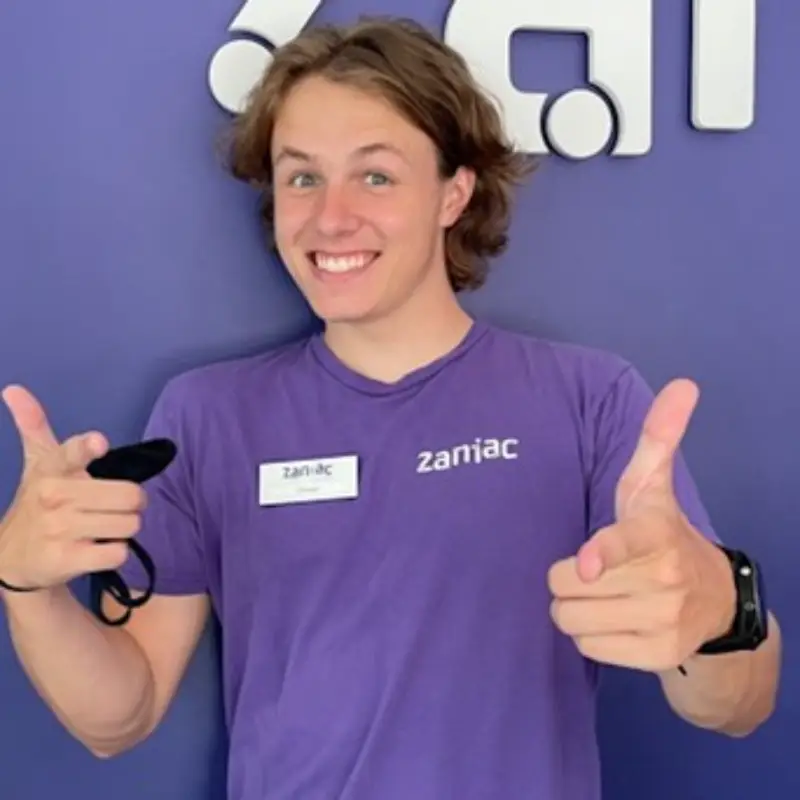 A young person in a purple Zaniac shirt smiles and points both index fingers at the camera, standing in front of a purple wall with a partial Zaniac logo. They wear a name tag and a smartwatch.