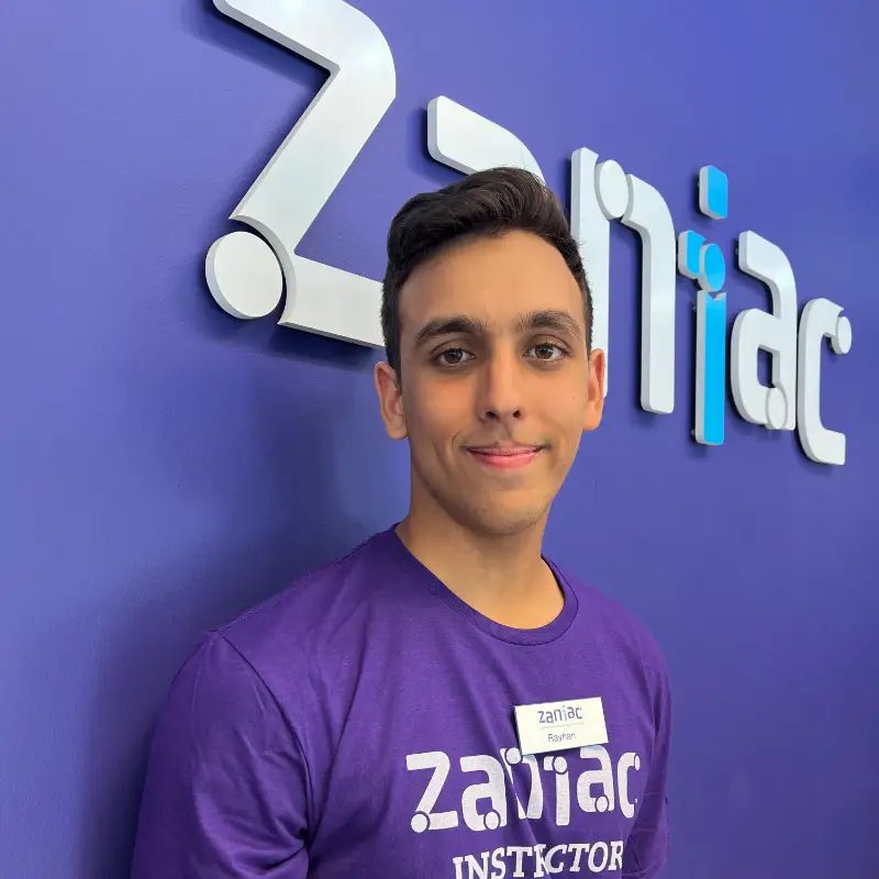 A young man wearing a purple "Zaniac INSTRUCTOR" shirt and a name tag stands in front of a purple wall with the Zaniac logo.