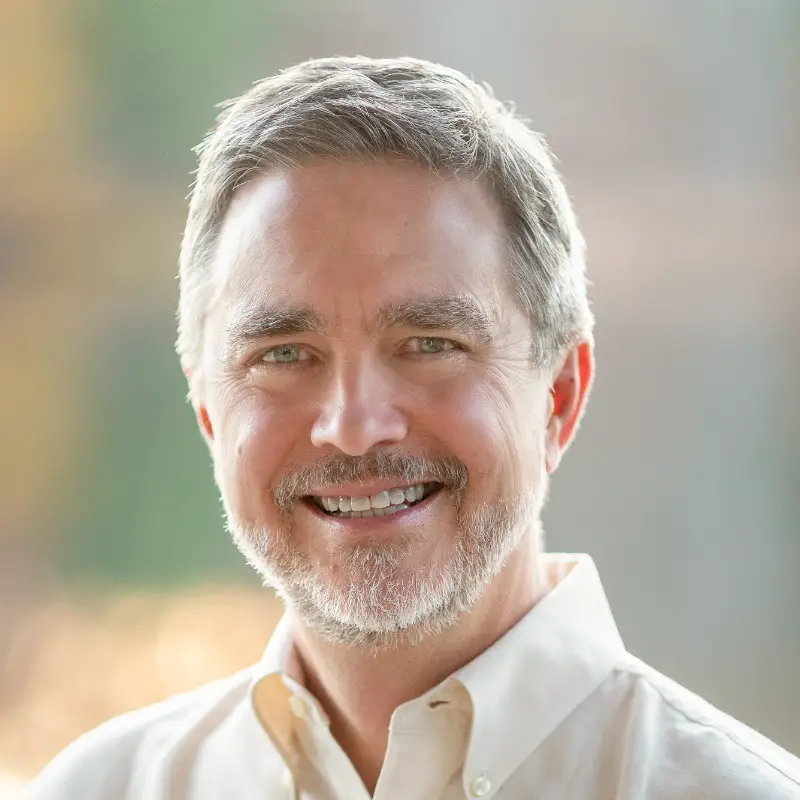 A middle-aged man with gray hair and a beard smiles warmly at the camera. He is wearing a light-colored button-up shirt, and the background is softly blurred with natural, outdoor lighting.