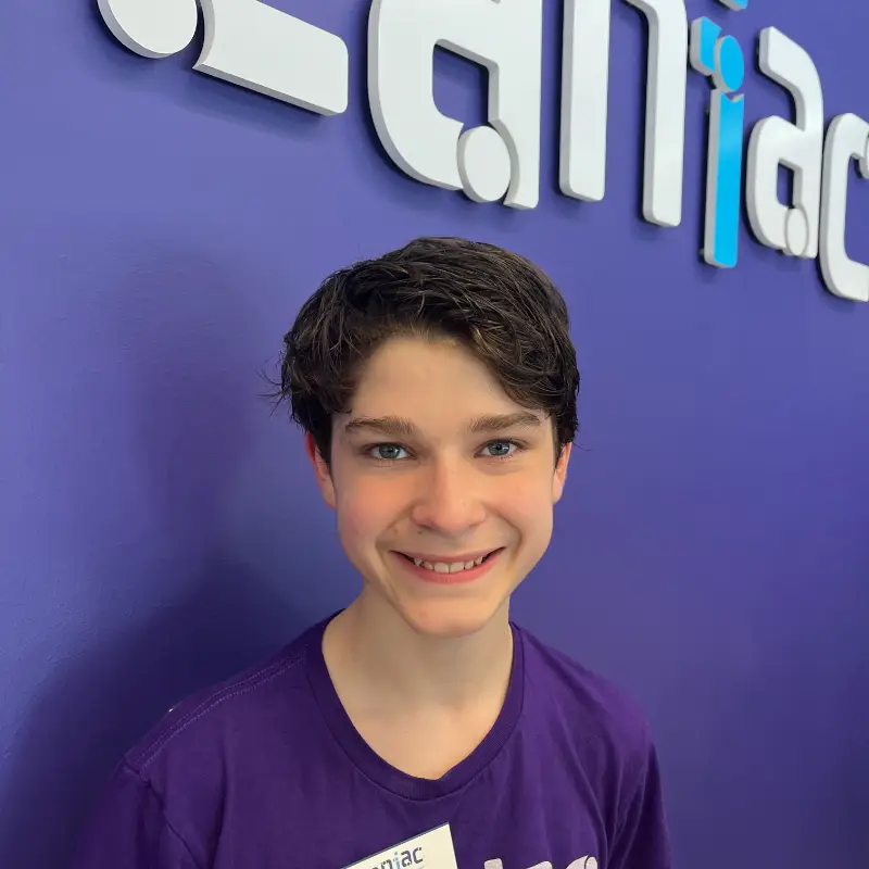 A smiling teenage boy with short brown hair stands in front of a purple wall with large white and blue letters. He is wearing a purple shirt and a name tag.