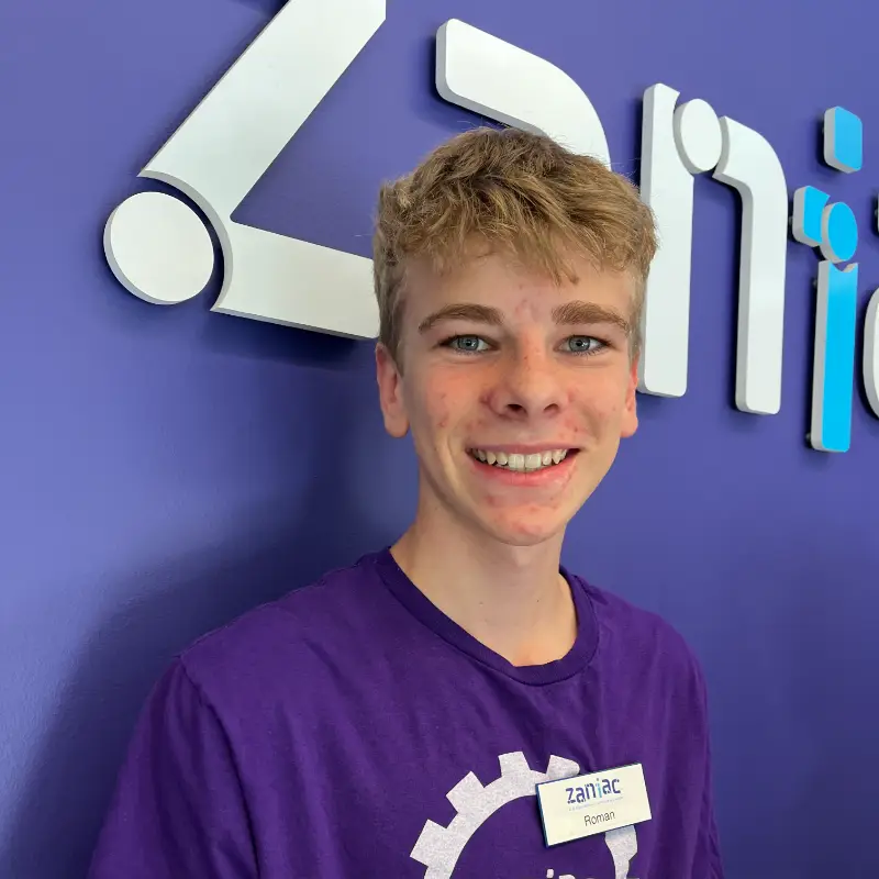 A teenage boy with blond hair and a purple T-shirt smiles at the camera. He is wearing a nametag and standing in front of a blue wall with a white and blue logo.