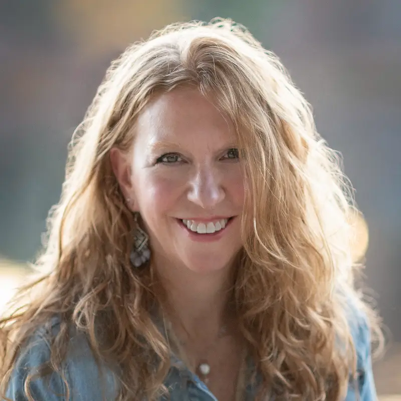 A woman with long, wavy, light brown hair smiles at the camera. She is wearing a denim shirt and dangling earrings, with a blurred outdoor background behind her.