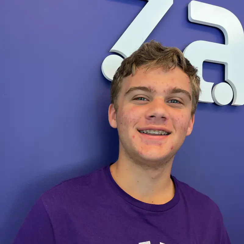 Teen boy with light brown hair and braces smiles in front of a purple wall with part of a white logo visible in the background. He is wearing a purple shirt.