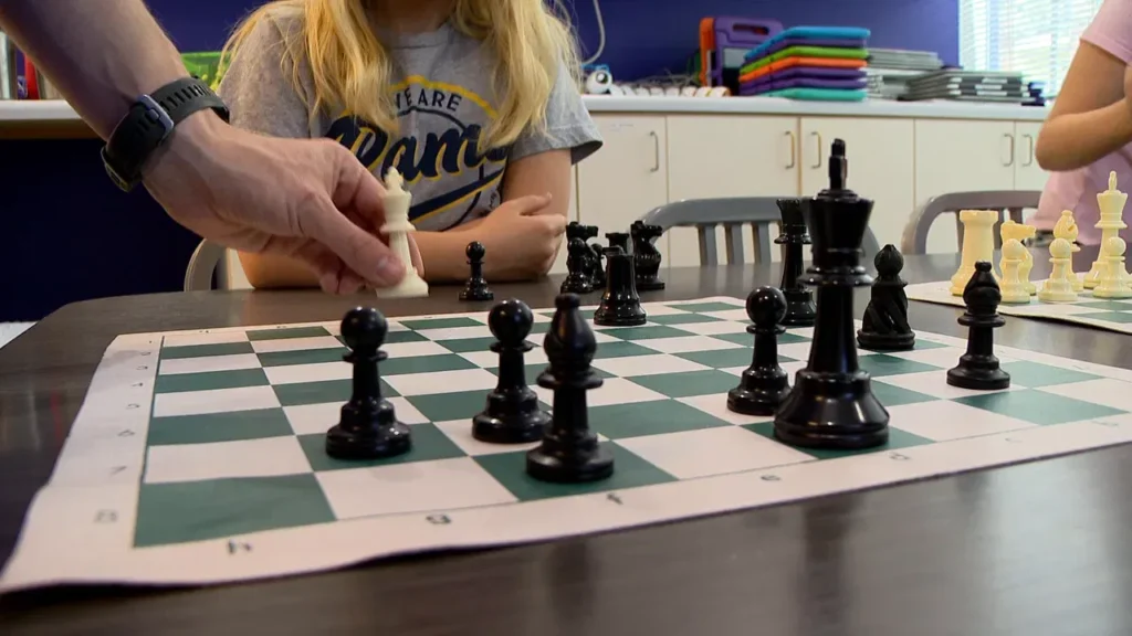 A person moves a white chess piece on a green and white chessboard, while a child with long blond hair watches. Stacked colorful trays and cabinets are visible in the background.