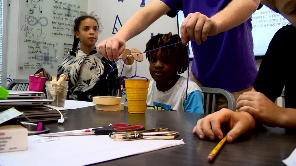 Three children are seated at a table working on a STEM project. One child is holding a string attached to a cardboard and cup contraption. The table is covered with papers, scissors, a pencil, and other materials.