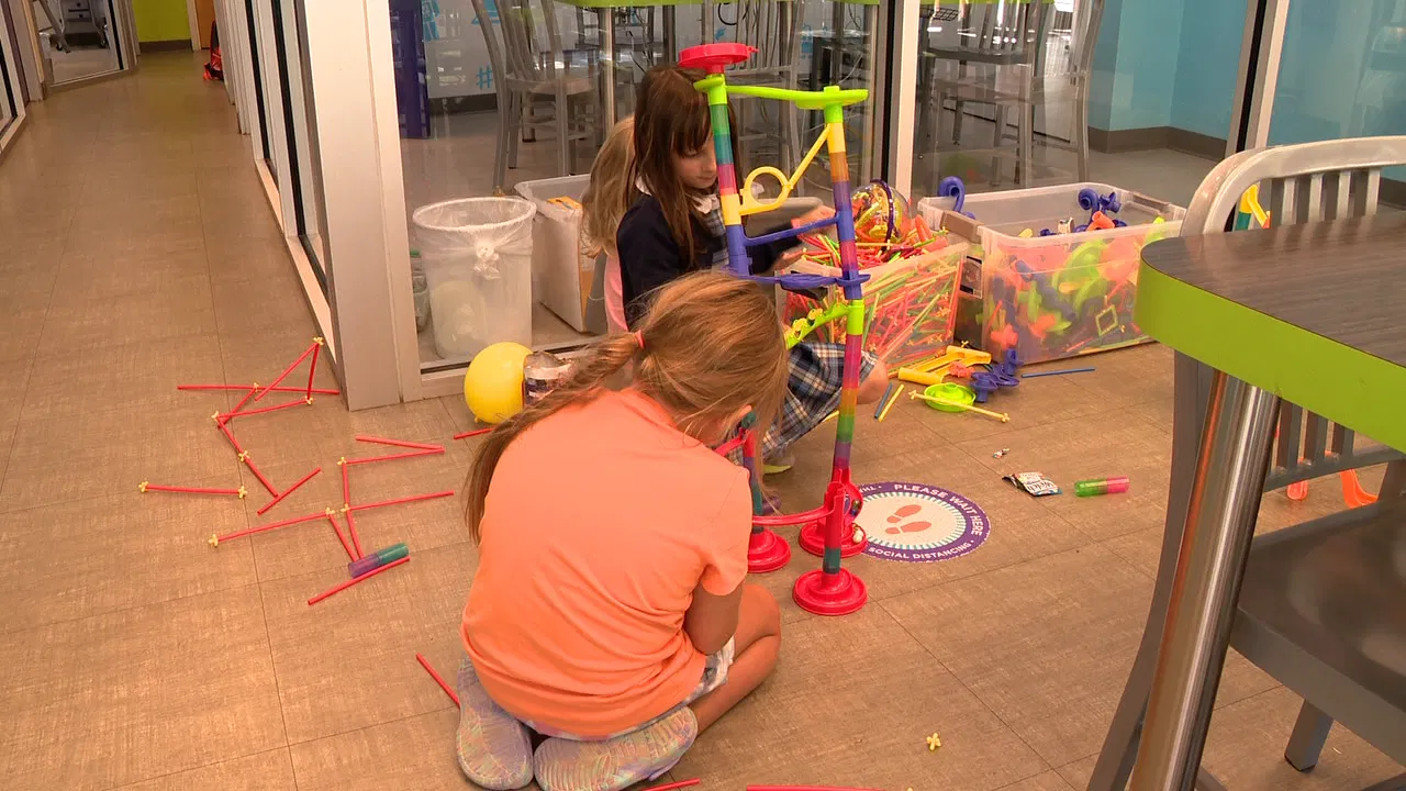 Two children sit on the floor building a colorful marble run with plastic pieces. Various toy parts are scattered around them. They are in a bright indoor space with glass walls and tables.