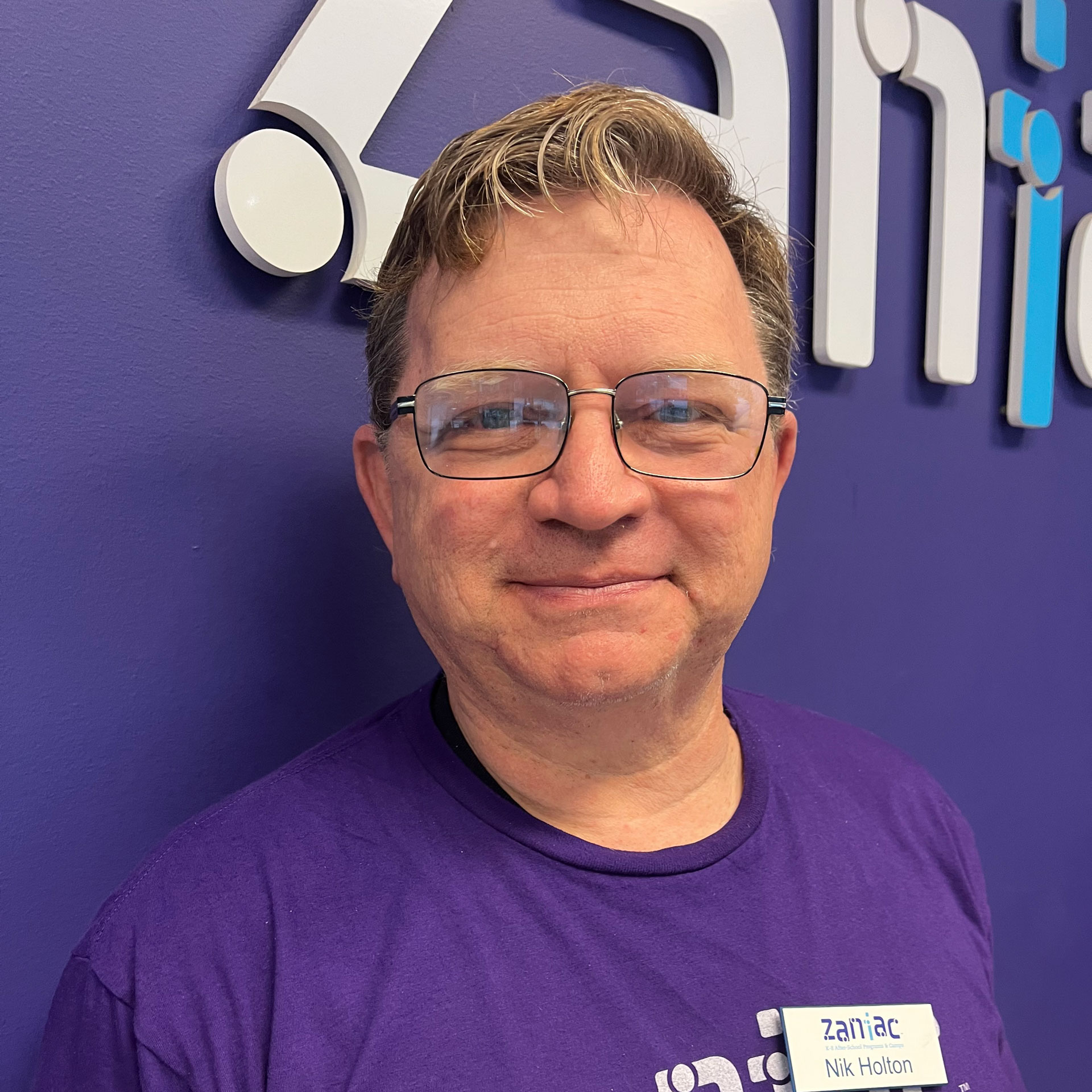 A man with glasses and short hair smiles while wearing a purple shirt and a name tag that says “Nik Holton” in front of a purple wall with the word “Zaniac” in large letters.